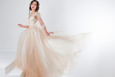 A teen girl with dark brown hair wearing a cream-colored formal dress poses on a white staircase with a white background.