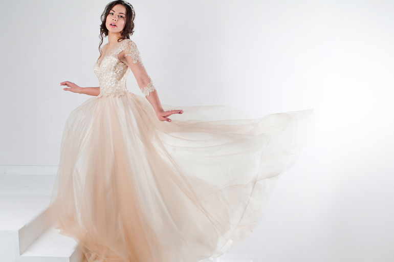 A teen girl with dark brown hair wearing a cream-colored formal dress poses on a white staircase with a white background.