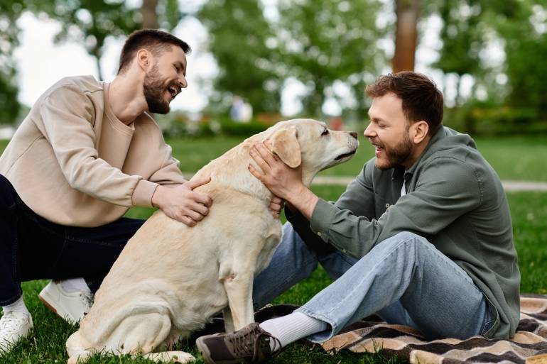 Two men laughing and smiling while hugging and touching a large white labrador in an outdoor environment with trees in background.