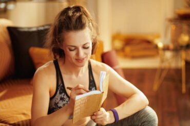 A woman wearing workout clothes smiles as she writes in a diary. She sits on the floor of a room in front of a couch.