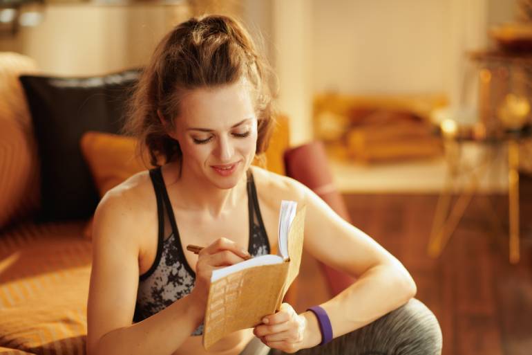 A woman wearing workout clothes smiles as she writes in a diary. She sits on the floor of a room in front of a couch.