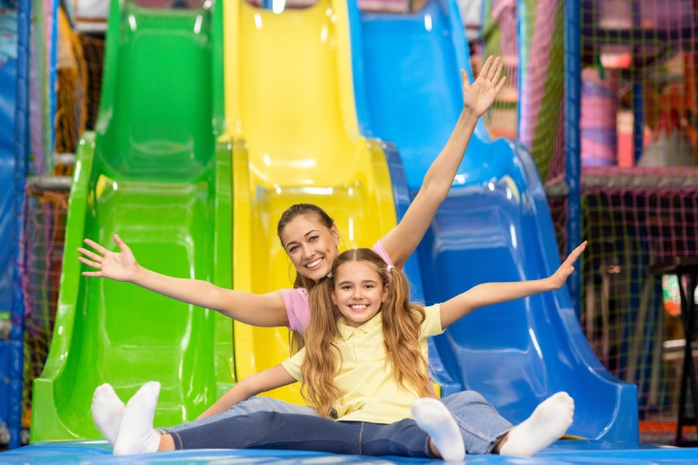 A happy mother and her daughter are riding down a colorful slide together at an indoor kids playground.