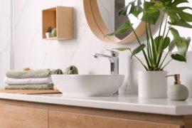 A wooden vanity has a white sink bowl sitting on top with a potted plant and green and white towels.