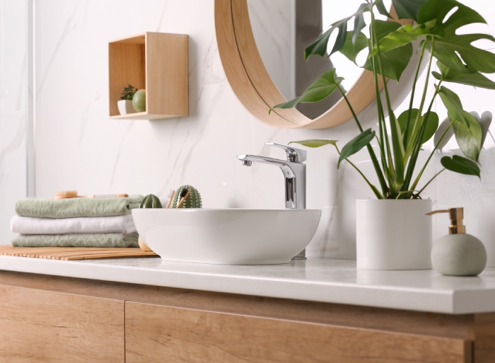 A wooden vanity has a white sink bowl sitting on top with a potted plant and green and white towels.