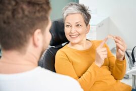 A woman sitting in a dentist's chair. She smiles and holds a dental implant while looking at a man in front of her.
