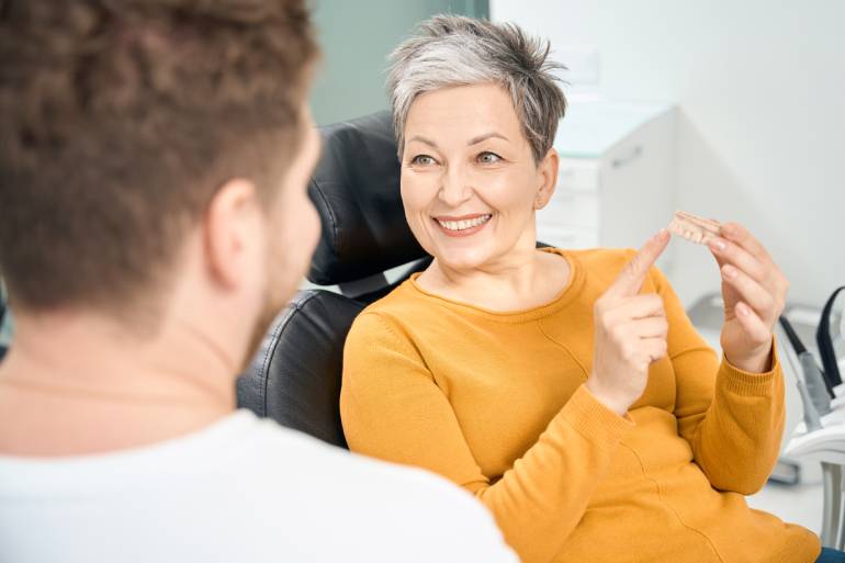 A woman sitting in a dentist's chair. She smiles and holds a dental implant while looking at a man in front of her.