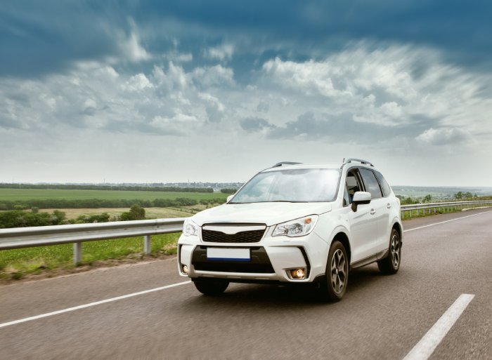 A passenger vehicle driving down a highway in the countryside. They SUV is white and there's a guardrail.