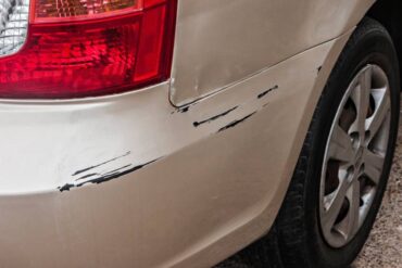 The rear-bumper of a beige car parked on epoxy flooring. The bumper has long black scratches from its side to the back.