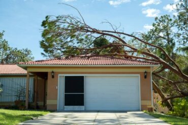A tree has collapsed onto the roof of a house's garage. The sky in the background is blue and mostly clear.