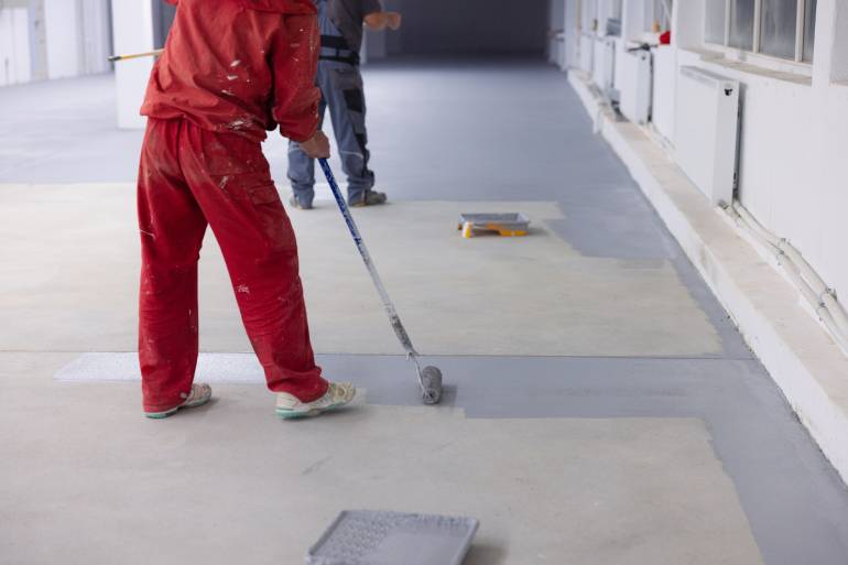 Two workers wearing protection uniforms applying acid stain to a large concrete floor indoors with paint rollers.