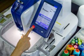 A man's hand pressing a red button below an embroidery machine's screen. The screen says "embroidery" on a white grid.