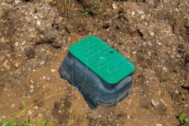A septic tank that is partially buried and surrounded by dirt and rocks. There is a green lid covering the tank.