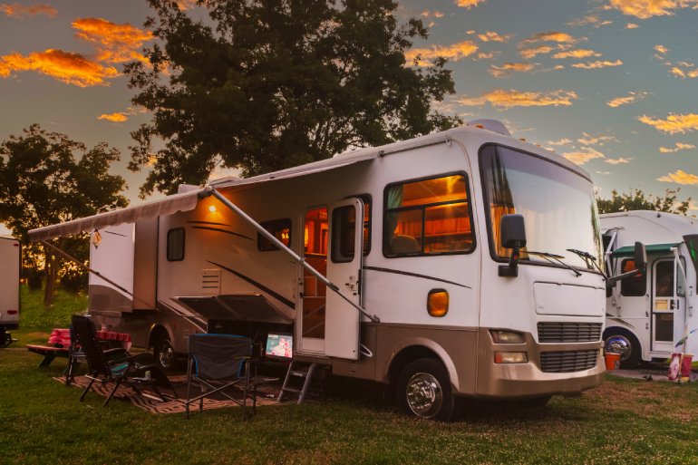 A large white and brown RV sits on the grass with the awning open and outdoor furniture set up. Another RV is next to it.