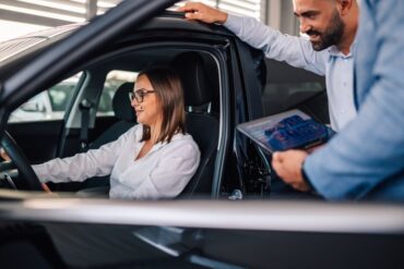 An automotive sales rep with a tablet, standing outside the vehicle with a car buyer sitting in the driver's seat.