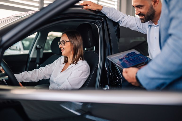 An automotive sales rep with a tablet, standing outside the vehicle with a car buyer sitting in the driver's seat.