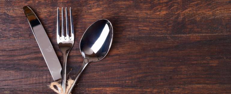 A wooden restaurant table with three pieces of silver cutlery wrapped in twine, including a knife, fork, and spoon.