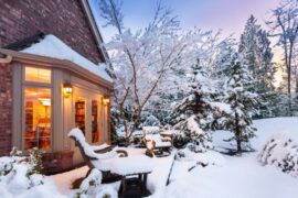 A home's backside and backyard are covered in snow while light floods outside through the back window going into a room.