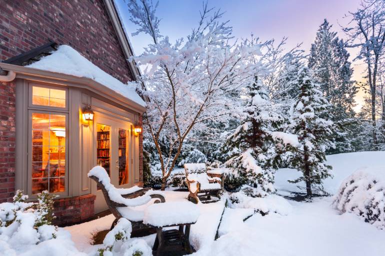 A home's backside and backyard are covered in snow while light floods outside through the back window going into a room.
