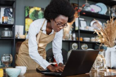 A Black female small business owner wearing an apron standing at her workstation. She's working on the laptop.