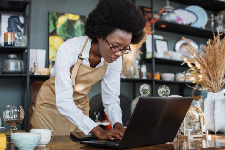 A Black female small business owner wearing an apron standing at her workstation. She's working on the laptop.