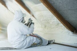 A professional spray foam insulation installer wearing a white protective suit and spraying the foam in between the wood.