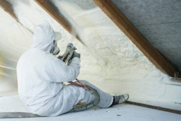 A professional spray foam insulation installer wearing a white protective suit and spraying the foam in between the wood.