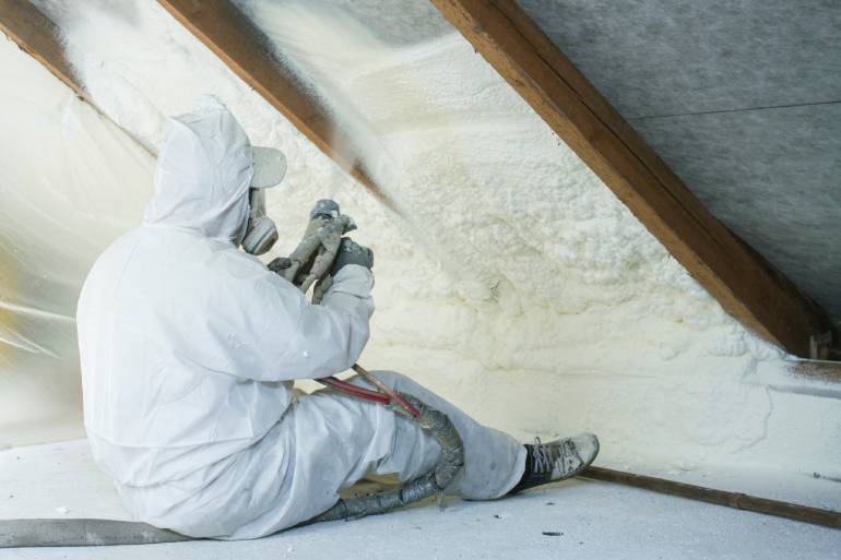 A professional spray foam insulation installer wearing a white protective suit and spraying the foam in between the wood.