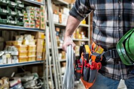 A worker in a hardware shop has his hand clenched around a roll of electrical wire. They also have a helmet and a tool belt.