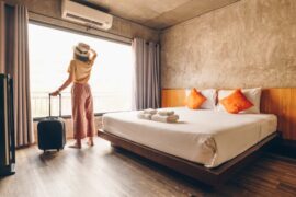A woman stands by the window in a hotel room with her hand on her hat and the other one holding her roller suitcase.