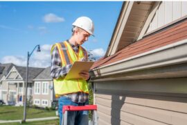 A roof inspector stands on a ladder and looks at shingles and a gutter. He writes down his observations on a clipboard.