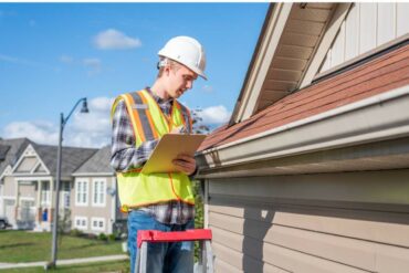 A roof inspector stands on a ladder and looks at shingles and a gutter. He writes down his observations on a clipboard.