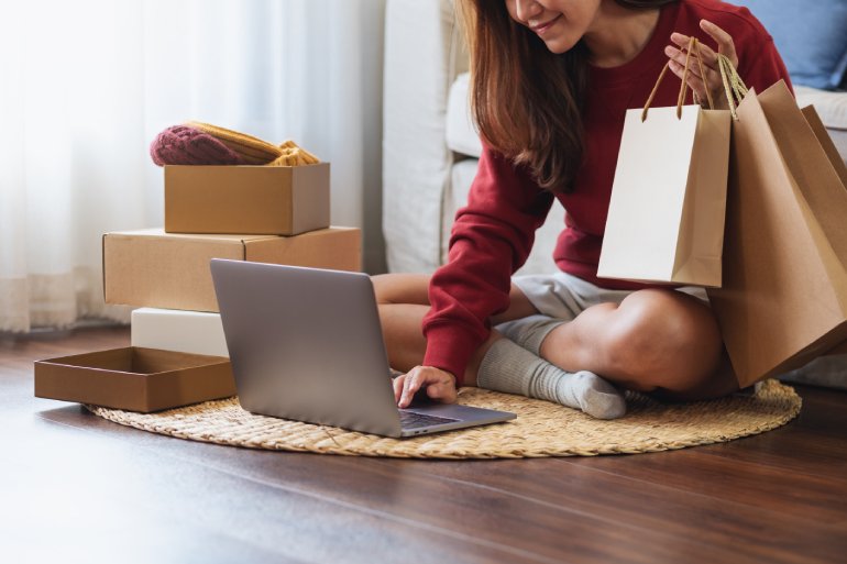 A woman sitting on the floor of her living room using her laptop. She is holding paper shopping bags in one hand.