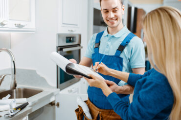 A woman signing a paper on a clipboard while a plumber in overalls smiles at her, holding the clipboard.