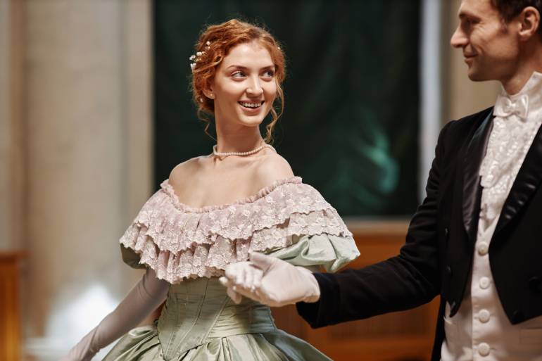 A woman in a delicate lace gown holding the hand of a gentleman in a suit and tie as they walk through a ballroom.