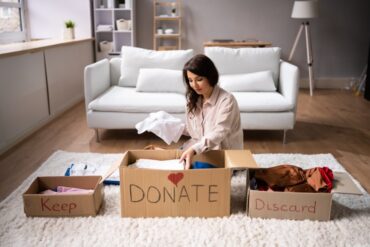 A woman sits on the floor of a living room and sorts items into boxes labeled “keep,” “donate,” and “discard.”