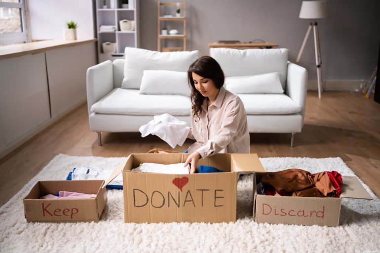 A woman sits on the floor of a living room and sorts items into boxes labeled “keep,” “donate,” and “discard.”