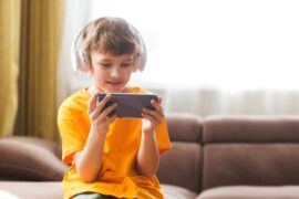 A young boy wearing a pair of white headphones while sitting on a couch and holding a smartphone in both hands.