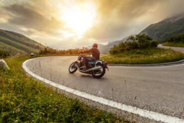 A person wearing a black leather jacket, jeans, and a black helmet rides a motorcycle on a curved road.