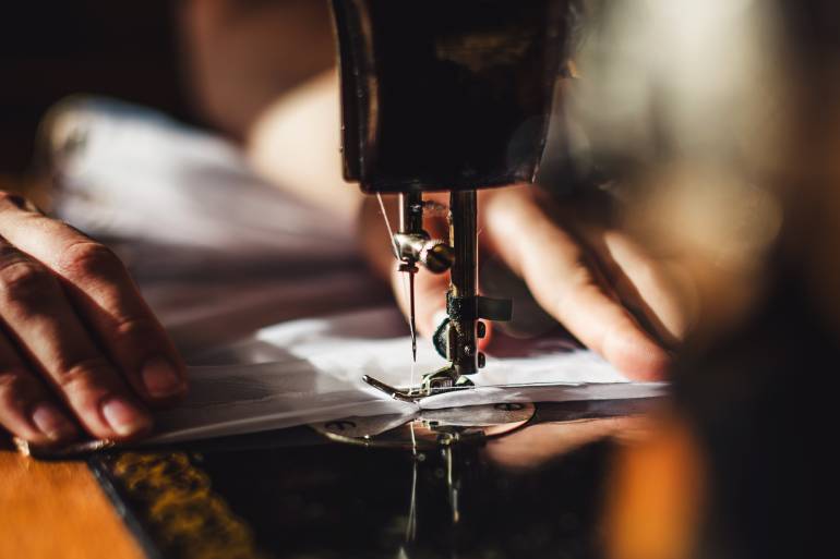 Close-up of two hands holding a white piece of fabric on an industrial sewing machine with white thread in needle.