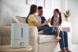 A white air purifier is sitting on a table and releasing vapor. A family of three is sitting on a couch in the background.
