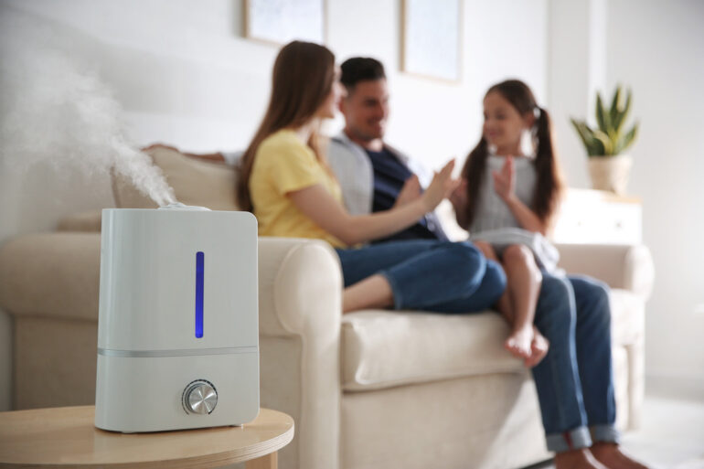 A white air purifier is sitting on a table and releasing vapor. A family of three is sitting on a couch in the background.