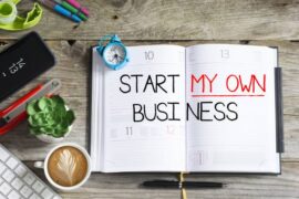 An open notebook planner with the words "start my own business" written on it. There are various items next to the planner.