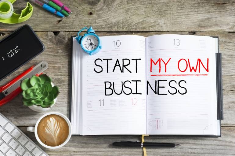 An open notebook planner with the words "start my own business" written on it. There are various items next to the planner.