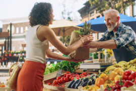 A male farmers market vendor smiles behind his fruits and vegetables stand as he hands a woman a bag of items.