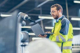 A factory worker stands next to a machine while holding a laptop. He's wearing a reflective jacket and glasses.