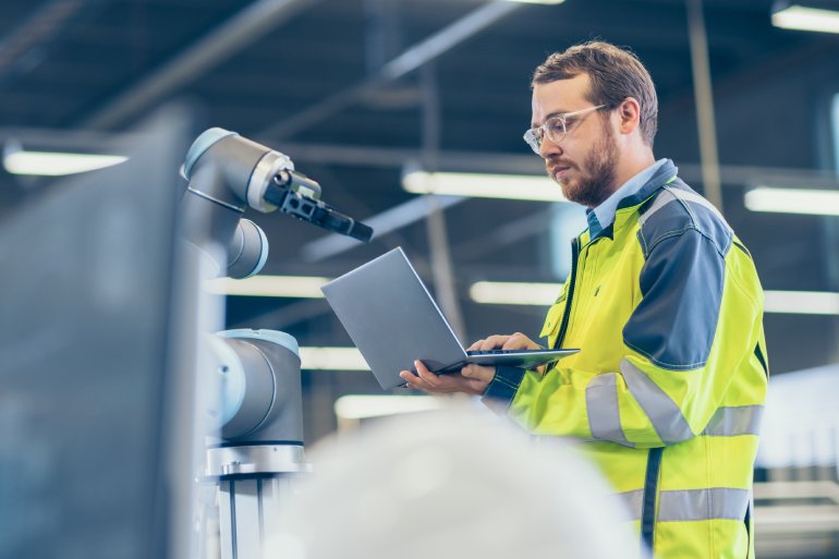 A factory worker stands next to a machine while holding a laptop. He's wearing a reflective jacket and glasses.