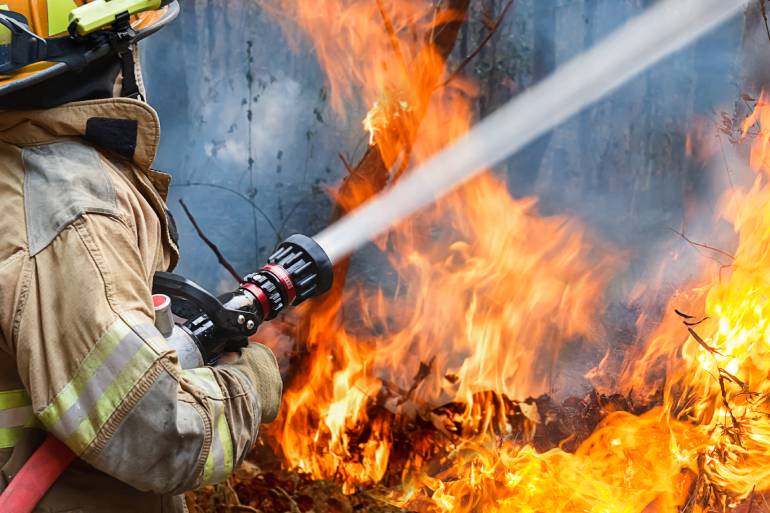 A close-up view shows a firefighter in a tan jacket and safety helmet spraying water from a red hose onto a wildfire.