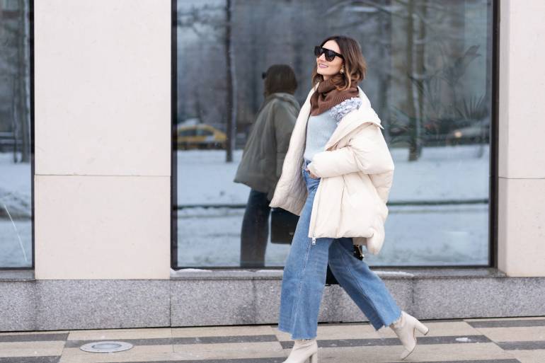 A woman walking on the sidewalk in a white puffer coat, cropped denim, and white heeled booties. She has sunglasses on.