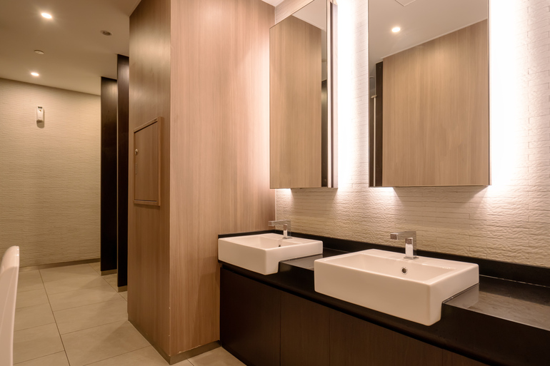 An ambient commercial bathroom with warm lights, large vanity mirrors, and basin sinks. There's a hand dryer to the left.