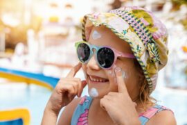 A young girl in a coloful hat and rainbow sunglasses smiling as she applies white sunblock on her cheeks.